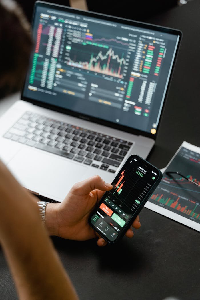 Person analyzing stock market data on a smartphone with a laptop displaying financial graphs in the background.